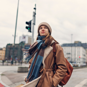 Eine junge Frau mit Mütze, Schal und Rucksack steht draußen in der Stadt und schaut nachdenklich zur Seite.