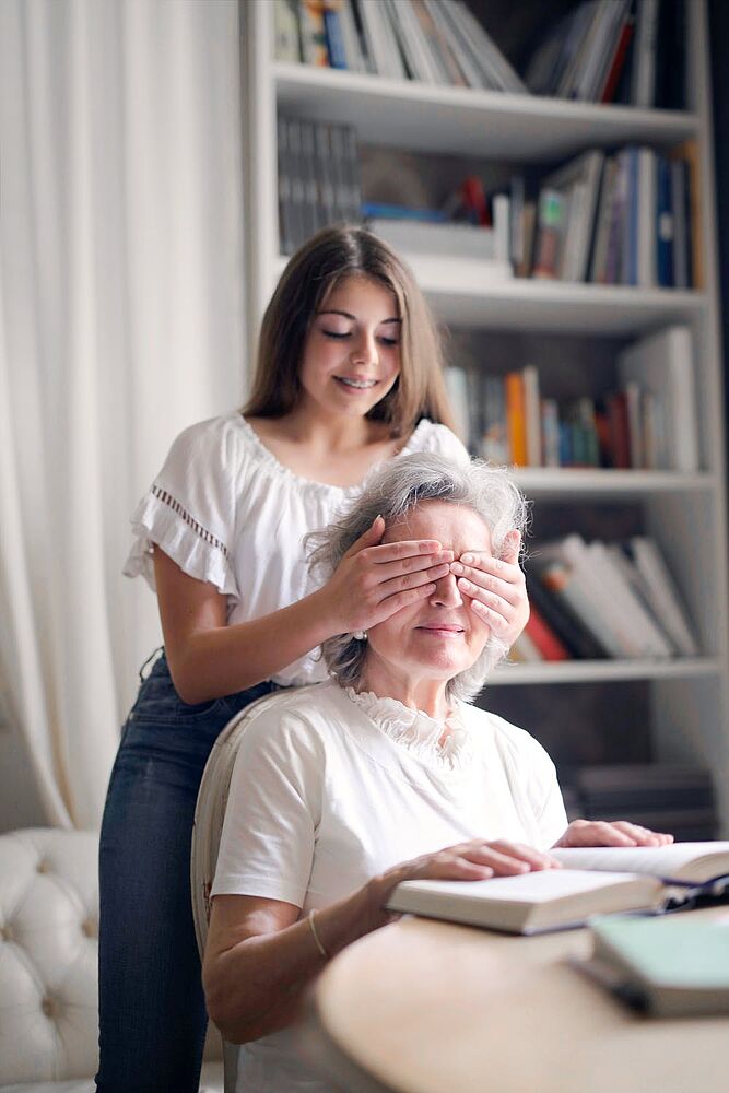 Ein Mädchen hält einer sitzenden Frau von hinten die Augen zu