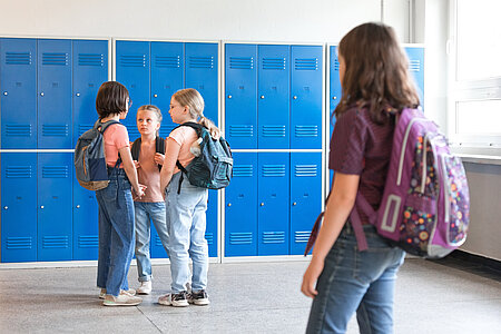 Vier Kinder stehen im Flur einer Schule. Ein Kind steht abseits der Gruppe und guckt traurig in ihre Richtung.